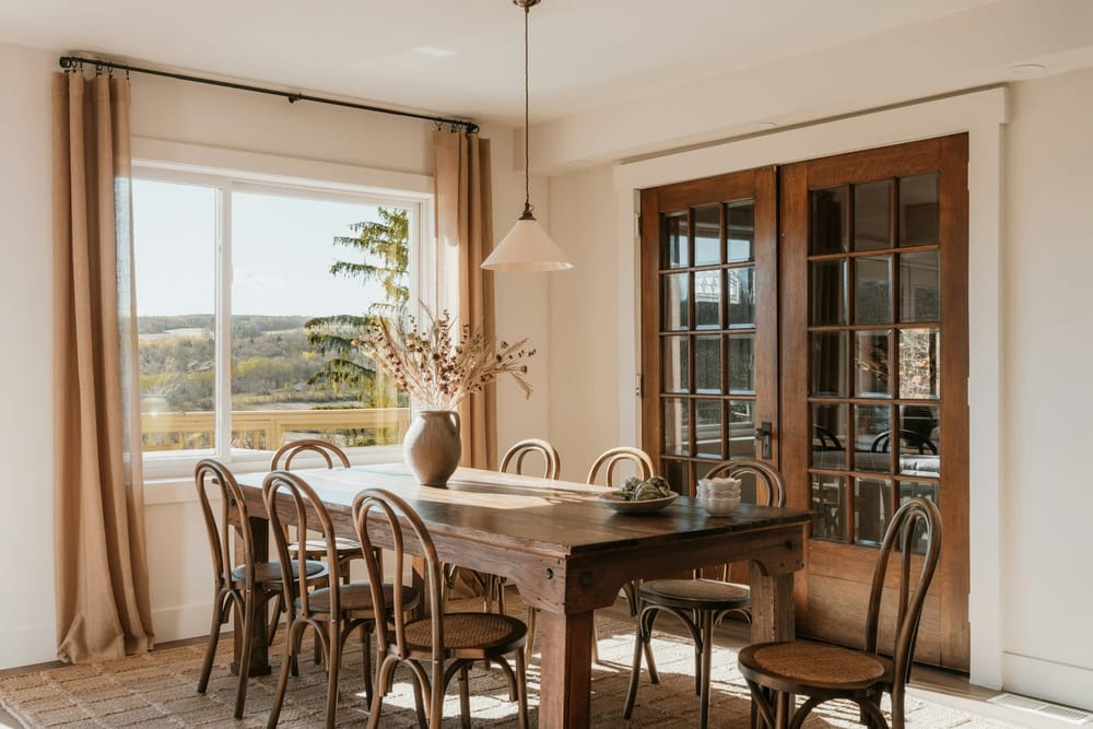 Een rustieke houten tafel op dikke houten poten, met stoelen van gebogen hout met een gevlochten zitting, een geweven kleed en een witte hanglamp