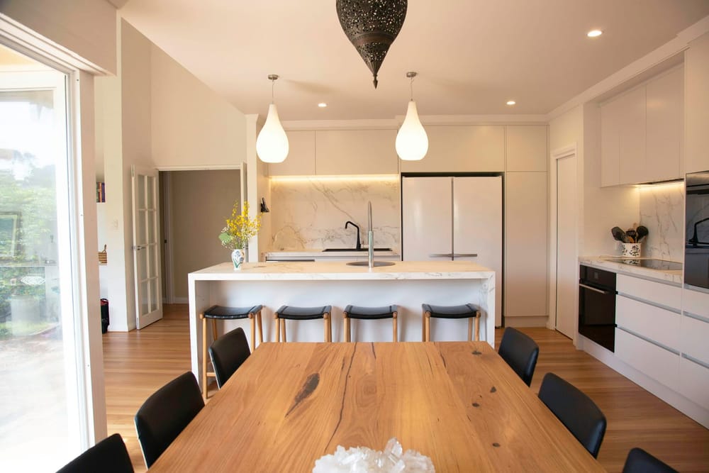 Een woonkeuken met een wit kookeiland met krukken en daarvoor een houten eettafel met zwarte stoelen