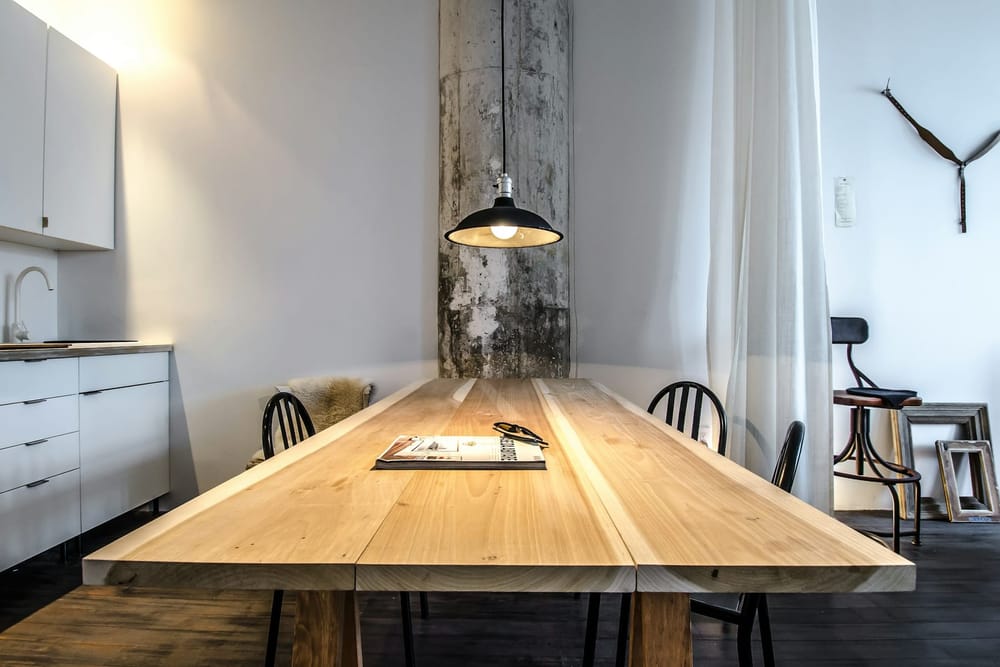 Een lange tafel van brede, lichte houten planken met zwarte stoelen en een zwarte hanglamp erboven