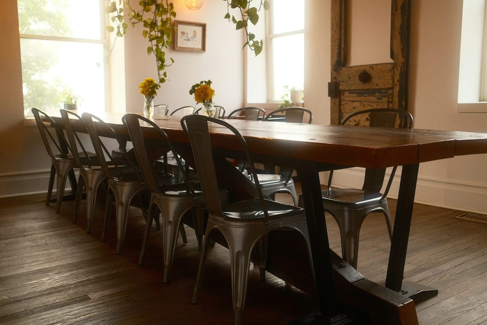 Een lange tafel met een dik, warmbruin houten blad op zwarte stalen poten, met grijze metalen stoelen op een donkere houten vloer