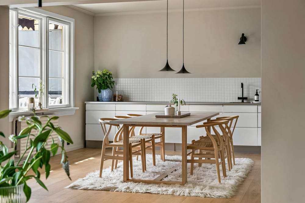 Een lichte houten tafel met lichte houten stoelen met een gevlochten zitting, een wit hoogpolig kleed en twee zwarte hanglampen in een witte keuken