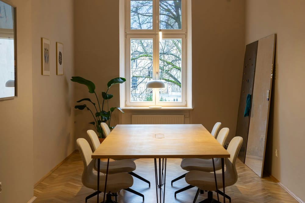 Een rechthoekige houten tafel op dunne zwarte stalen poten, met een klepje in het midden van het blad en zes witte stoelen op een draaivoet, voor een raam