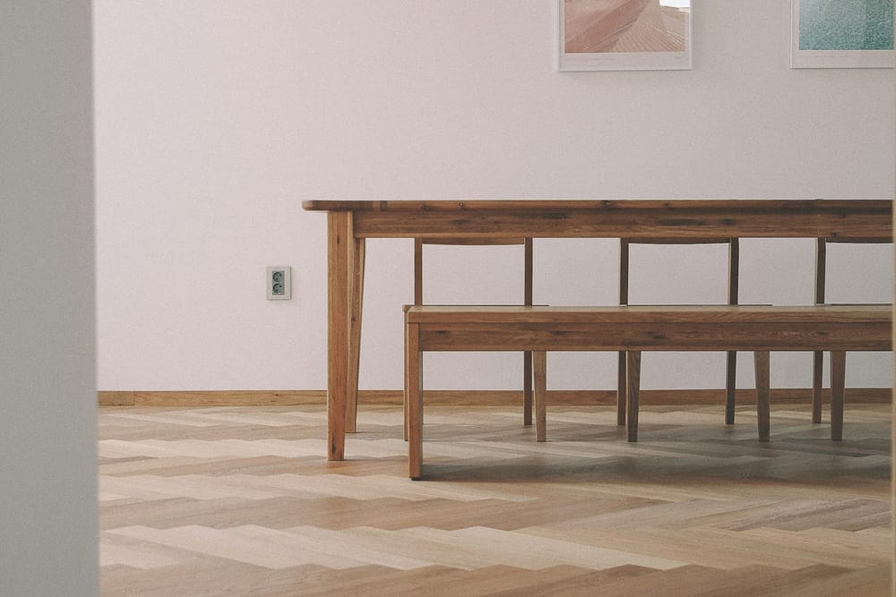 Een houten eettafel van opzij, met een brede rand onder het blad, een bank ervoor en stoelen erachter